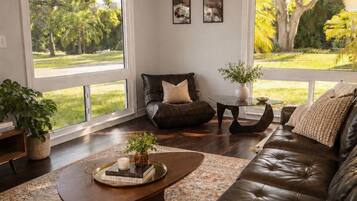 A living room with a leather sofa, a coffee table, a ceiling fan, and large windows with a view of greenery.