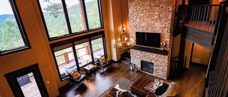 A spacious living room with a stone fireplace, large windows, and a high ceiling.