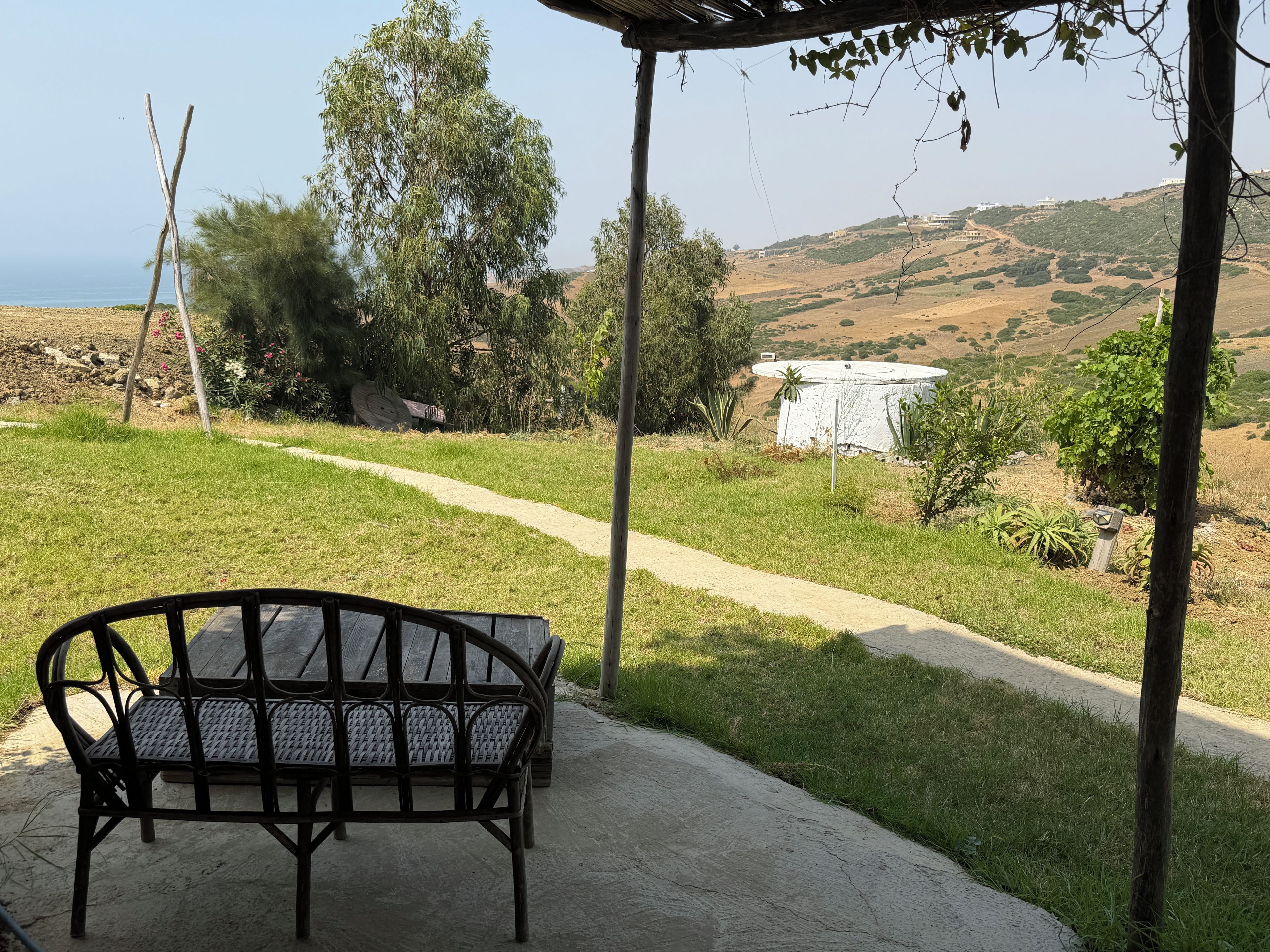 Un espacio de estar al aire libre con techo, una banca, un sendero de concreto y vistas a un paisaje montañoso con vegetación y un edificio a lo lejos.