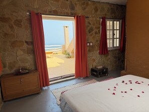 A room with a stone wall, a bed with rose petals, a wooden dresser, and a view of the sea through a window. - nilamane restaurant & atlantic view (Sahel Chamali)
