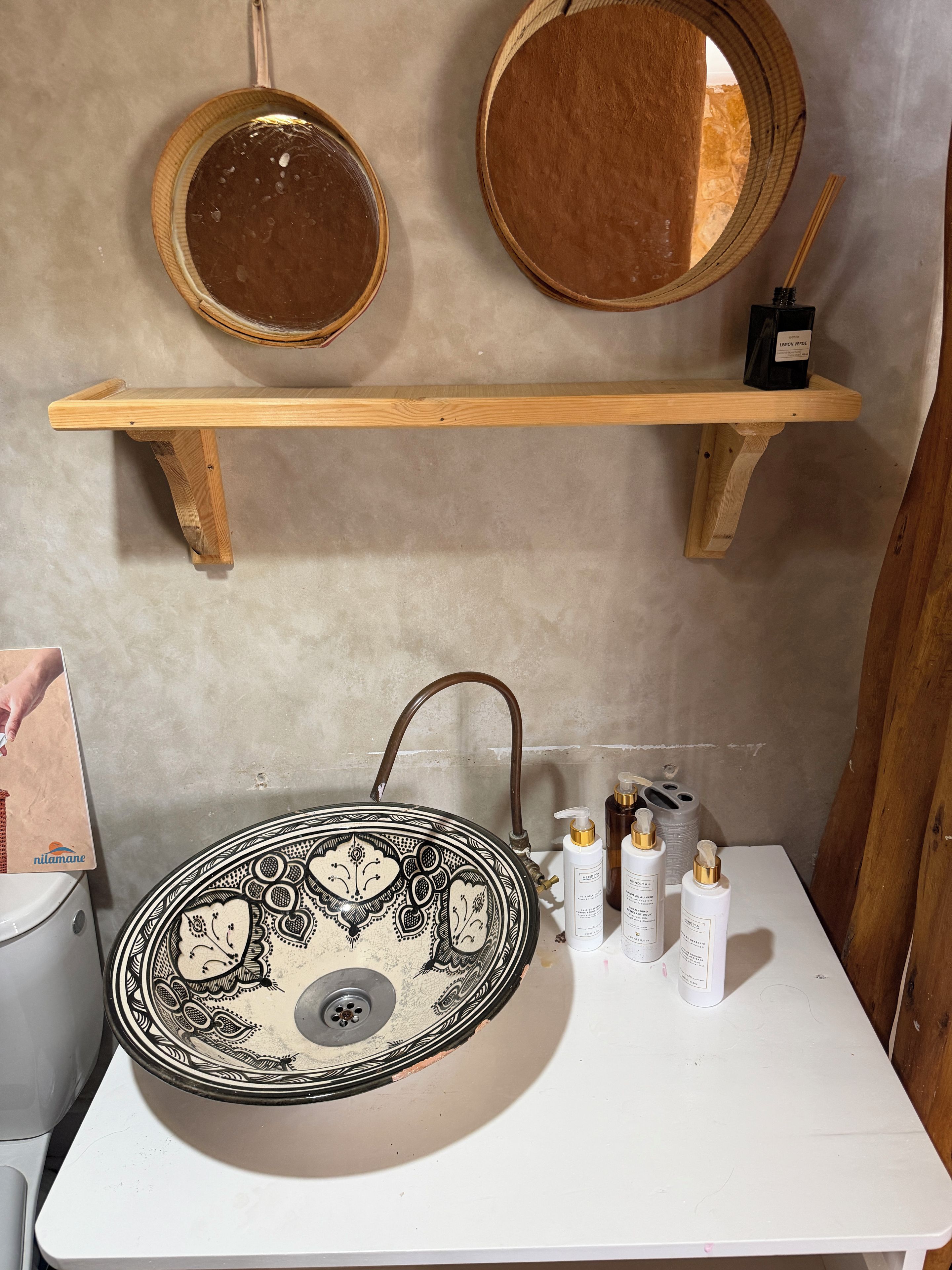 A bathroom with a round sink, two mirrors, a shelf with a bottle, and toiletries.