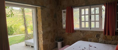 A bedroom with stone walls, a bed with a white bedspread, red curtains, and a view of a garden through a window.