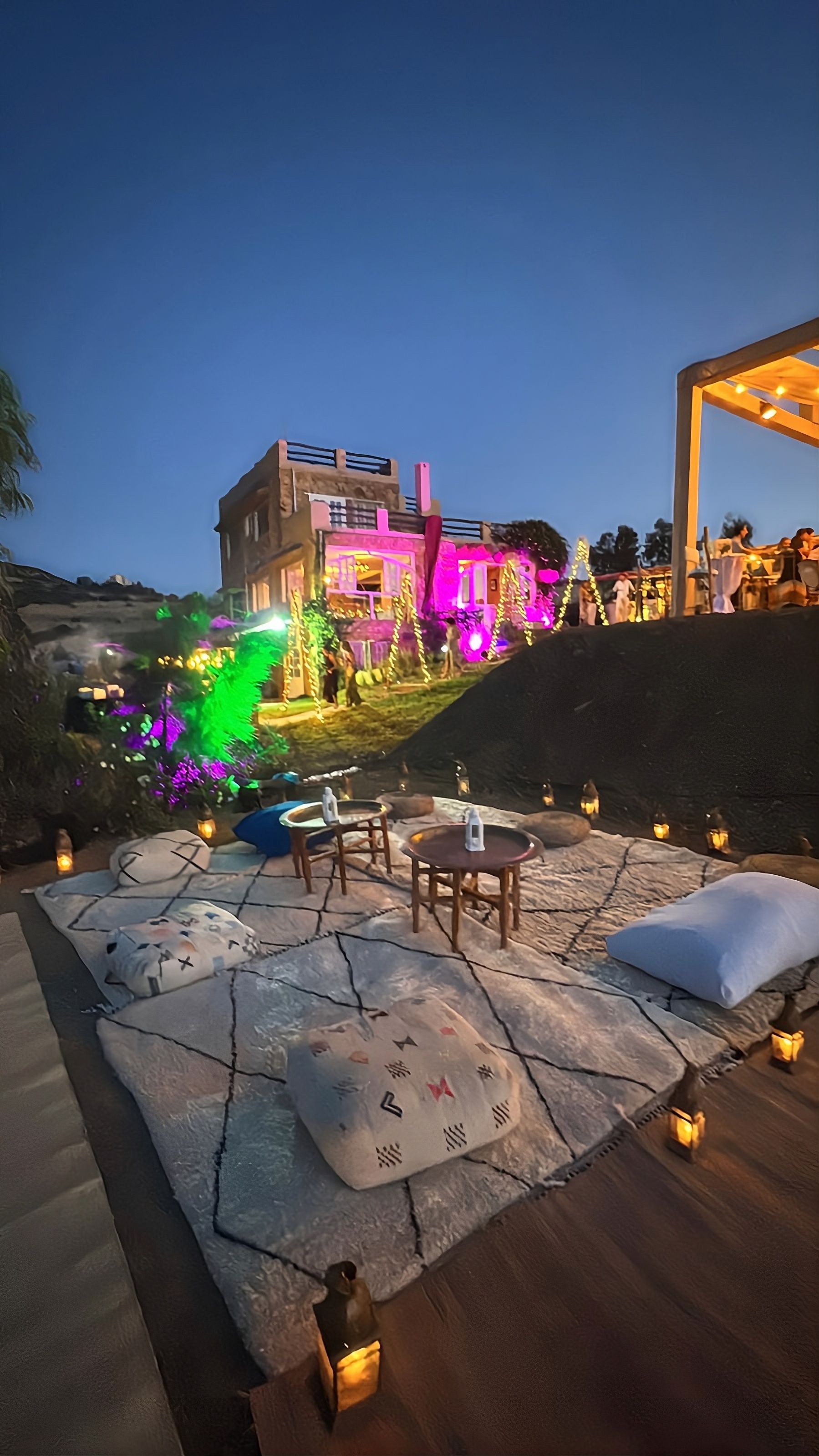 An outdoor seating area with stone flooring and tables, surrounded by decorative lighting and a view of a building with illuminated windows.