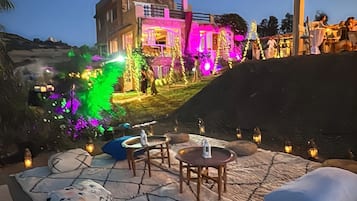 An outdoor seating area with stone flooring and tables, surrounded by decorative lighting and a view of a building with illuminated windows.