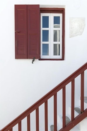 A white wall with a window featuring red shutters, a white door, and a wooden staircase railing. - Alana Pension (Mykonos)