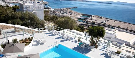Zona de piscina con sillones de descanso y sombrillas, vista al mar y un pueblo lejano.