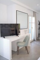 Una sala de estar moderna con una televisión de pantalla plana sobre una consola blanca, un sillón azul claro, un espejo grande en la pared y una ventana con cortinas.