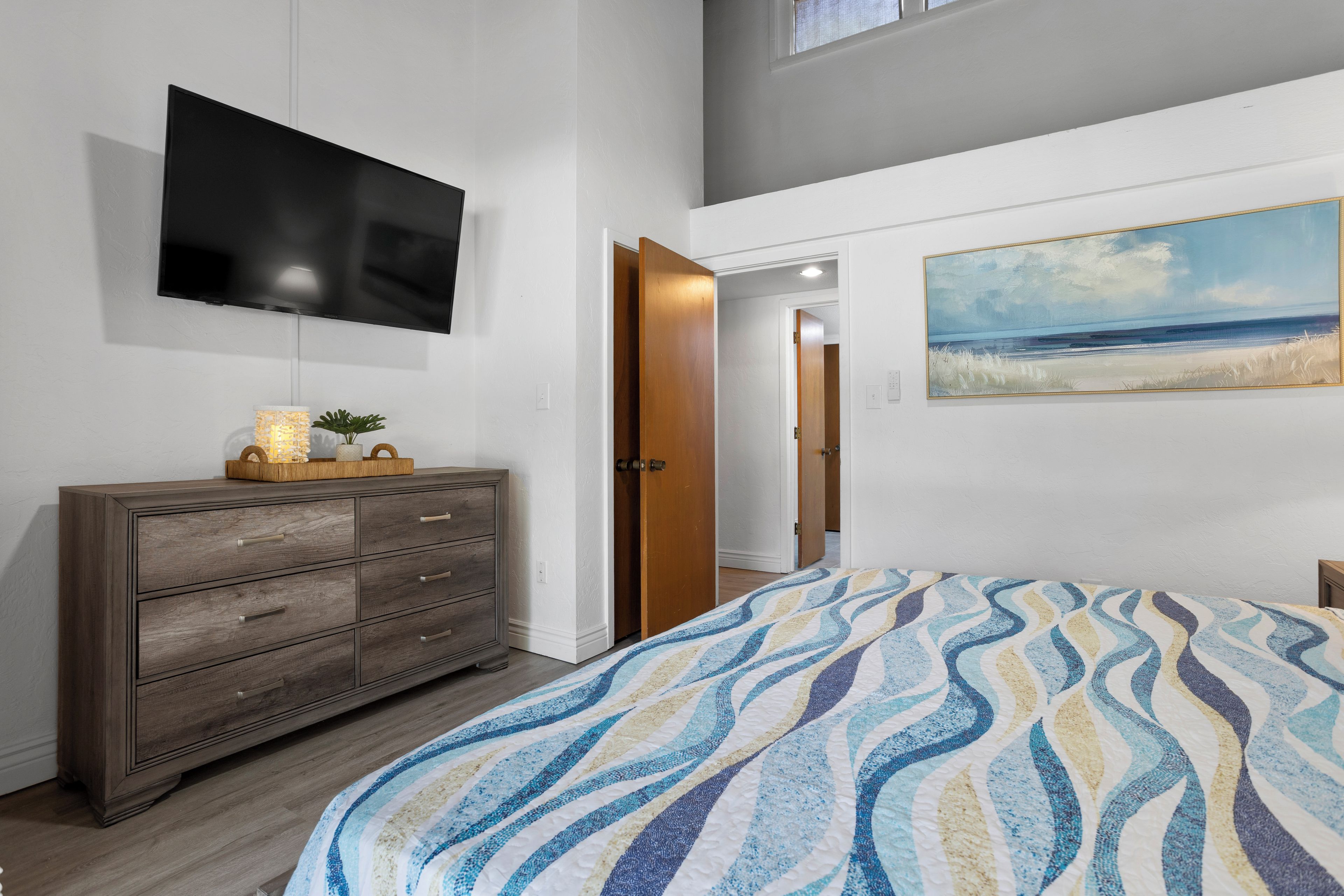 Une chambre à coucher avec un lit, un téléviseur fixé au mur, une commode en bois et un tableau représentant une scène de plage.