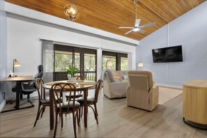 A spacious living room with a wooden ceiling, a round dining table with chairs, a sofa, a TV mounted on the wall, and large windows offering a view of greenery outside. - 657 Olde Camelot Circle (Haines City)