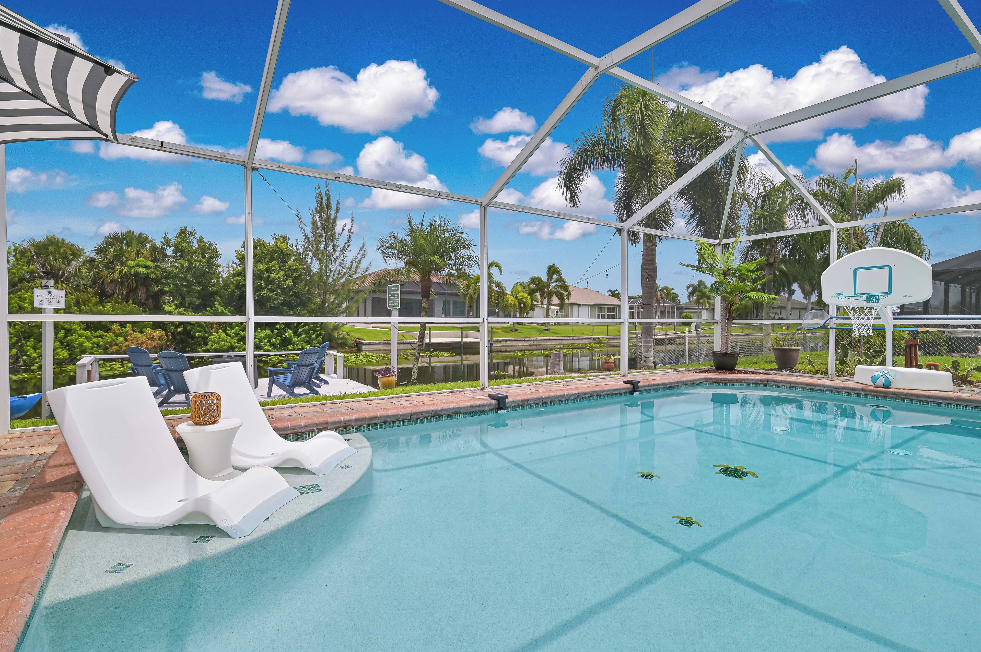 A swimming pool with lounge chairs and a basketball hoop, surrounded by palm trees and a clear sky.