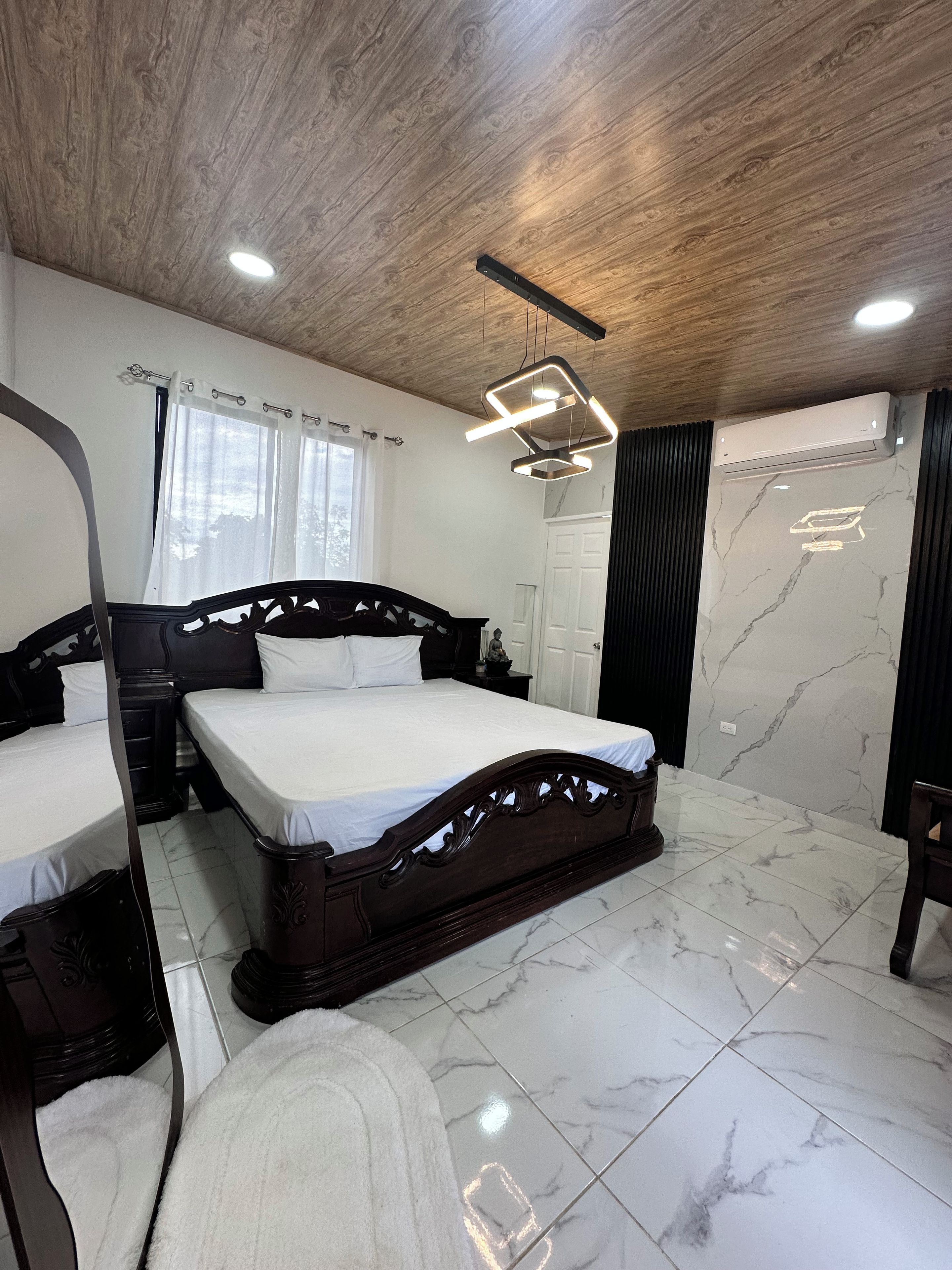 Une chambre avec un plafond en bois, un lit avec une tête de lit, une table de chevet, un miroir, une fenêtre avec des rideaux et un appareil de climatisation.
