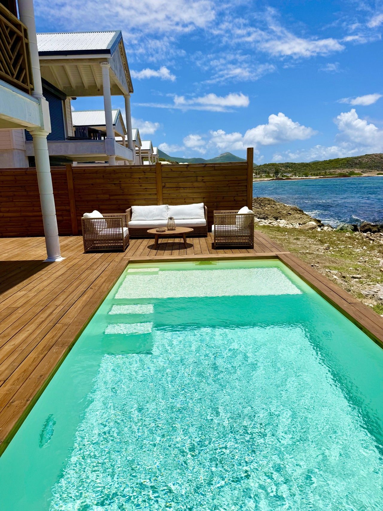A swimming pool with a wooden deck, two wicker chairs, and a white sofa.