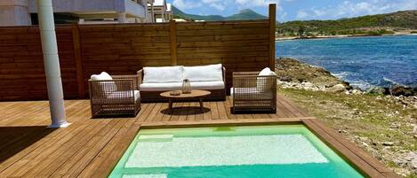 A swimming pool with a wooden deck, two wicker chairs, and a white sofa.