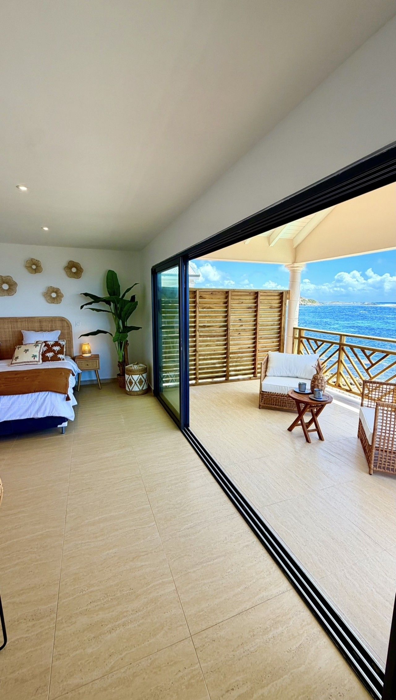 Una habitación amplia con una cama, una mesita de noche con lámpara, una planta grande y un balcón con muebles de mimbre.