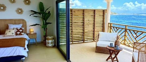 Una habitación amplia con una cama, una mesita de noche con lámpara, una planta grande y un balcón con muebles de mimbre.