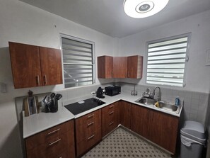 A compact kitchen with wooden cabinets, a black countertop, a sink, and a window with blinds. - Cozy 1-bedroom apartment in beautiful San Juan with AC (San Juan)