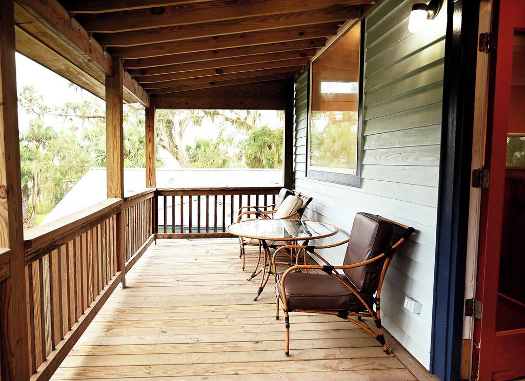 A wooden deck with a glass table and chairs, a red door, and a view of greenery.