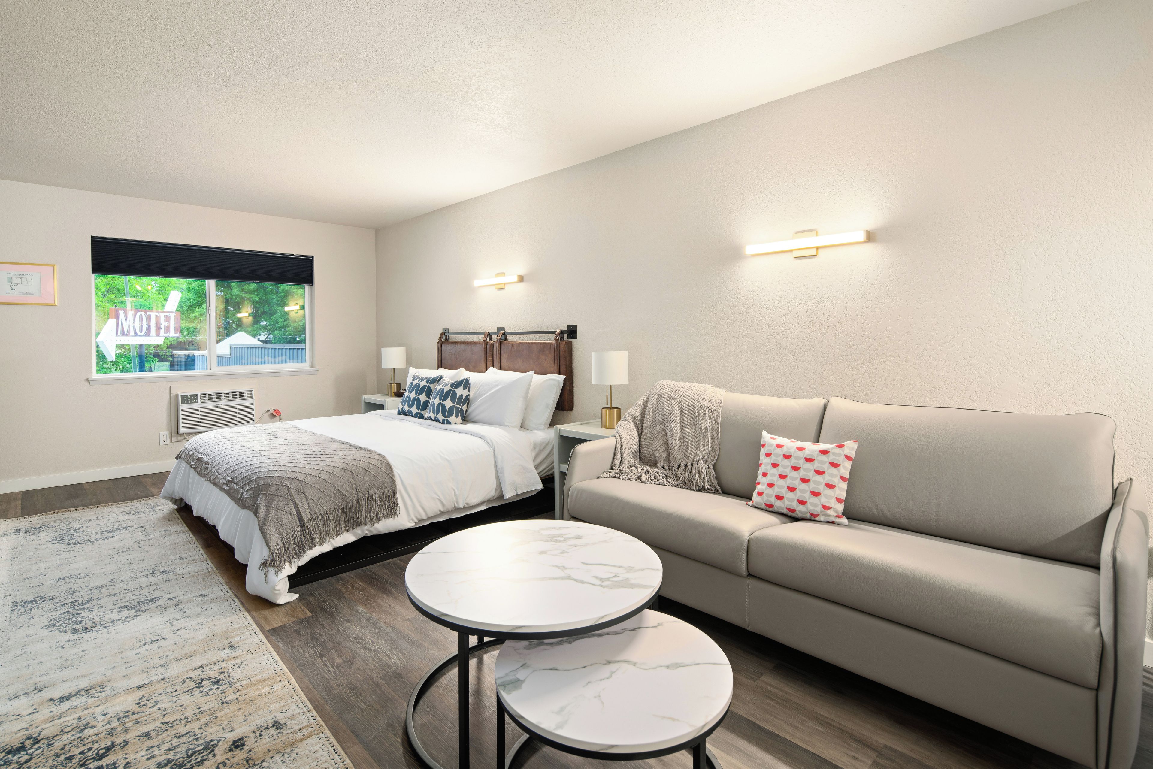 A modern bedroom with a bed, a sofa, a small table, and a window with a view of a motel sign.
