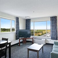 A hotel room with a flat-screen TV, a dining table, a sofa, and large windows offering a view of the landscape.