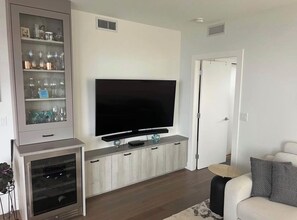 A living room with a flat-screen TV mounted on a wooden cabinet, a glass cabinet with decorative items, and a sofa with cushions. - White Rock Ocean View 2 Bedroom Furnished Condo / November to April (White Rock)