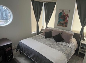 A bedroom with a bed, curtains, a window, a painting, and a desk. - White Rock Ocean View 2 Bedroom Furnished Condo / November to April (White Rock)