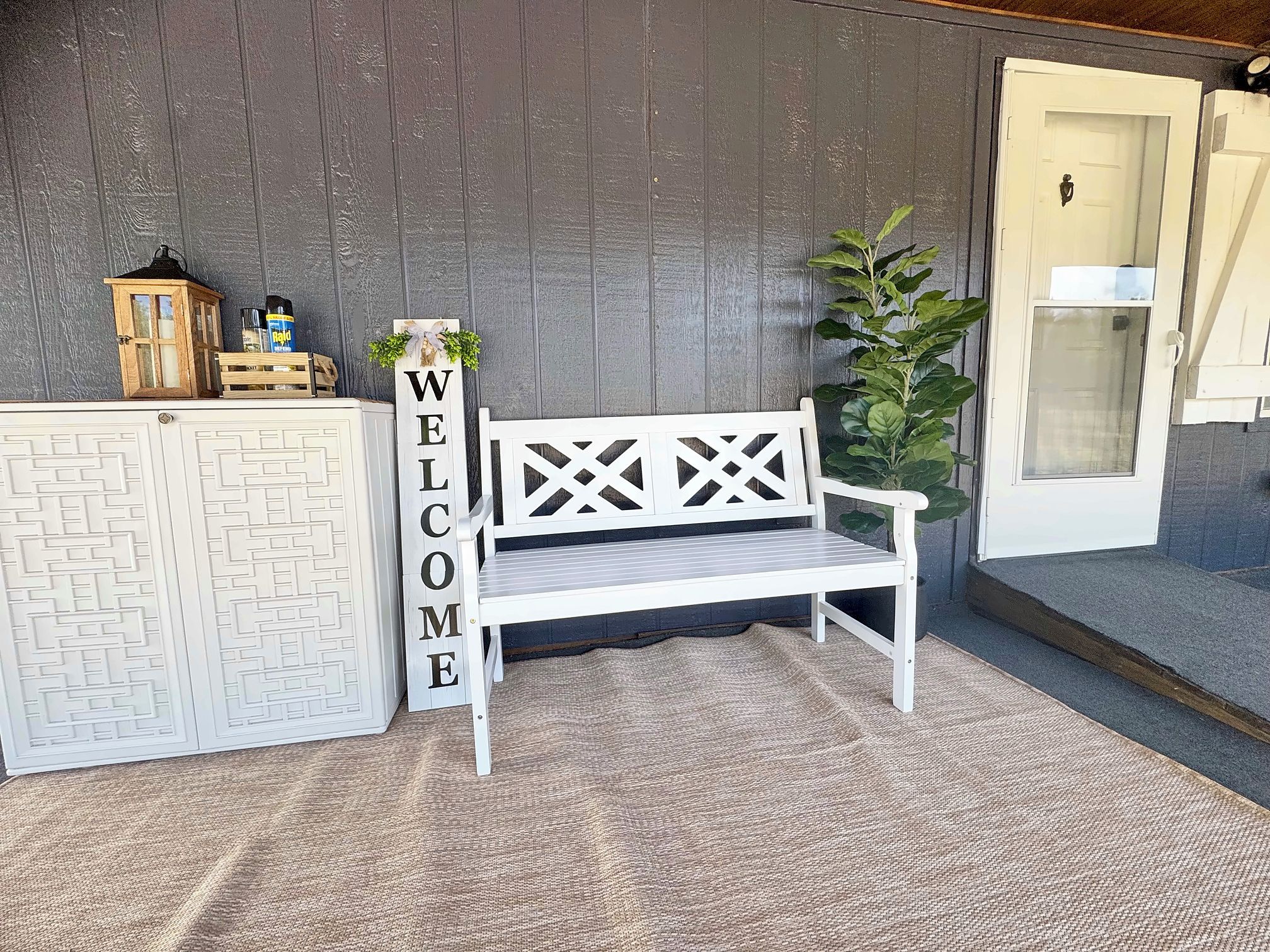 A white bench with a lattice backrest, a 'WELCOME' sign, a wooden crate with items, and a door with a glass panel.