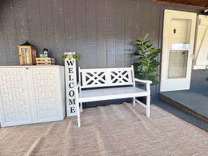 A white bench with a lattice backrest, a 'WELCOME' sign, a wooden crate with items, and a door with a glass panel.
