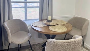 A room with a round table, two chairs, a window with curtains, and a view of an outdoor area.