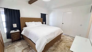 A bedroom with a bed, a bedside table, a lamp, a window with curtains, and a door.