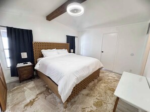 Ein Schlafzimmer mit Bett, Nachttisch, Lampe, Fenster mit Vorhängen und einer Tür.
