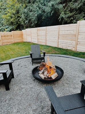 A circular fire pit with logs, surrounded by a gravel area, with a wooden fence and greenery in the background. - Whispering Cottage (Bothell)