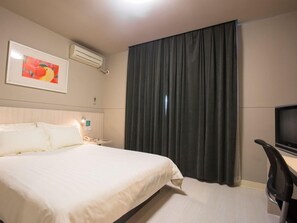 Habitación de hotel con cama, escritorio, silla, televisión, aire acondicionado, un cuadro en la pared y ventana con cortinas.