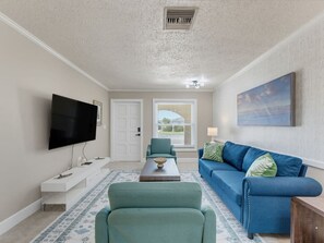 Uma sala de estar com televisão de tela plana, um sofá azul, uma poltrona verde e uma mesa de centro.