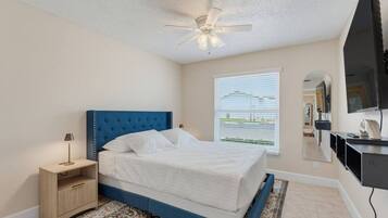 A bedroom with a bed, a nightstand, a ceiling fan, a window with blinds, and a flat-screen TV mounted on the wall.