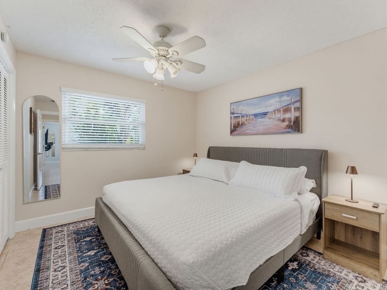 A bedroom with a bed, a ceiling fan, a nightstand, a lamp, a window with blinds, and a painting on the wall.