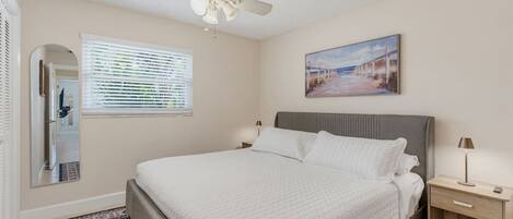 A bedroom with a bed, a ceiling fan, a nightstand, a lamp, a window with blinds, and a painting on the wall.