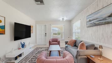A living room with a flat-screen TV, a sofa, a pink ottoman, and a coffee table.