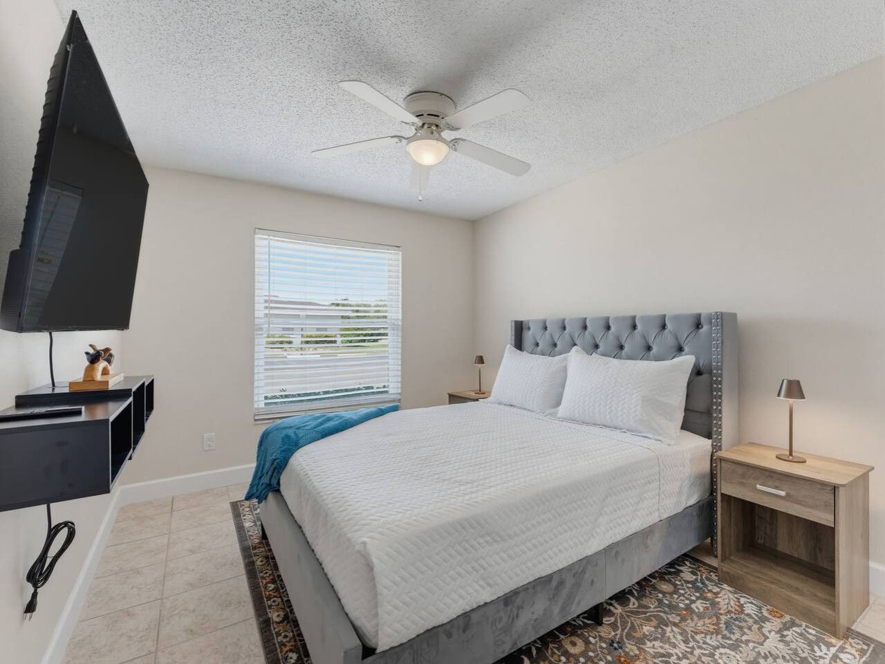 A bedroom with a bed, a ceiling fan, a TV mounted on the wall, a window with blinds, and a nightstand with a lamp.