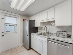 A compact kitchen with white cabinets, a stainless steel refrigerator, a built-in dishwasher, and a toaster. - Saltwater & Bubbles 2 | Indian Rocks Beach (Indian Rocks Beach)