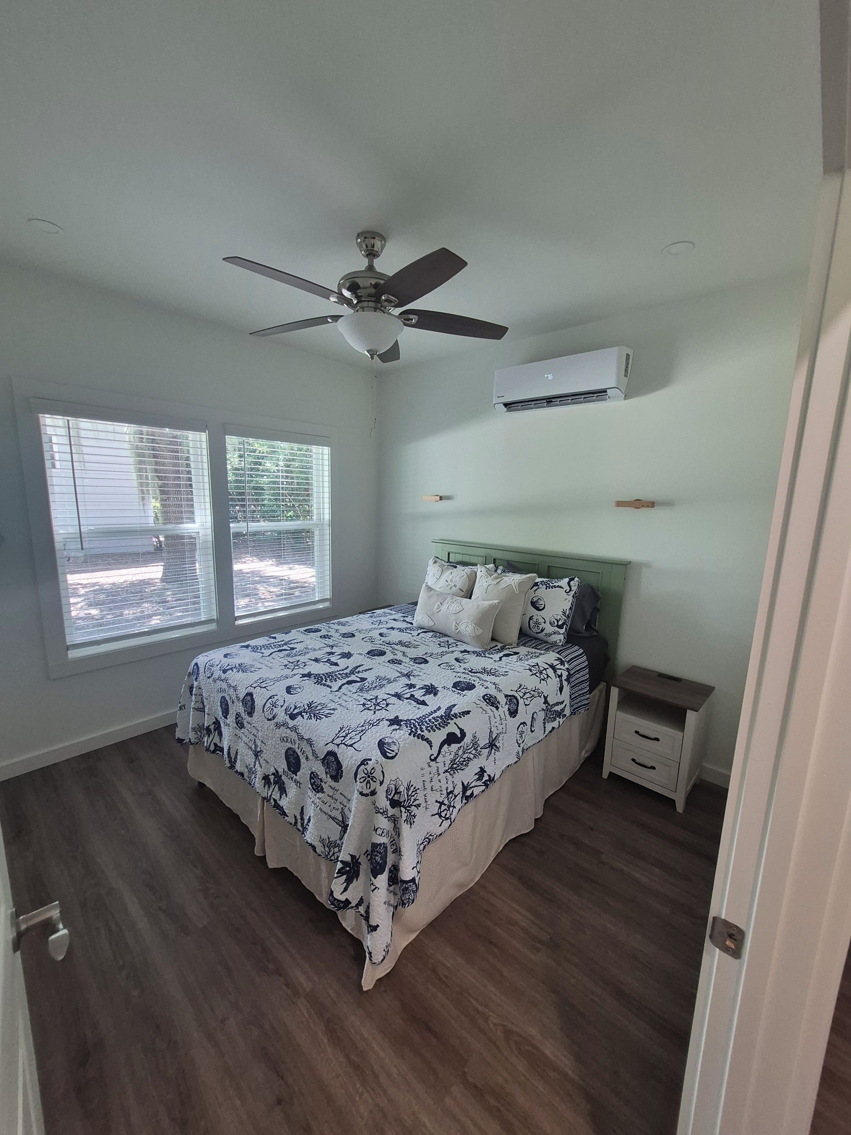 A bedroom with a bed, a ceiling fan, an air conditioning unit, a nightstand, and a window with blinds.