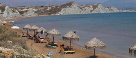 A beach with sun loungers and thatched umbrellas, a clear sky, and distant mountains.