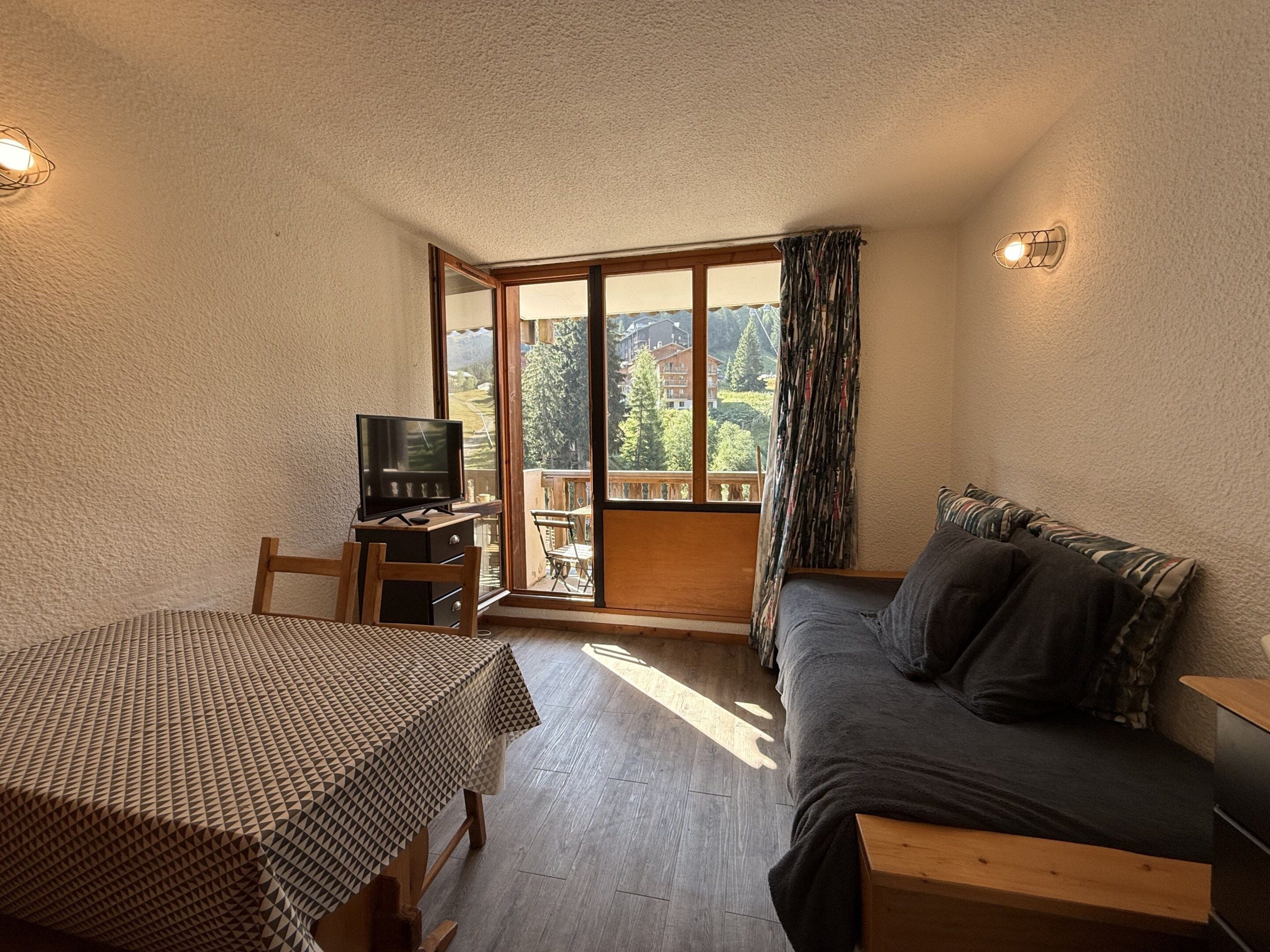Quarto com mesa de madeira, sofá, televisão e porta de vidro deslizante que dá para área externa com vista para árvores e edifícios.