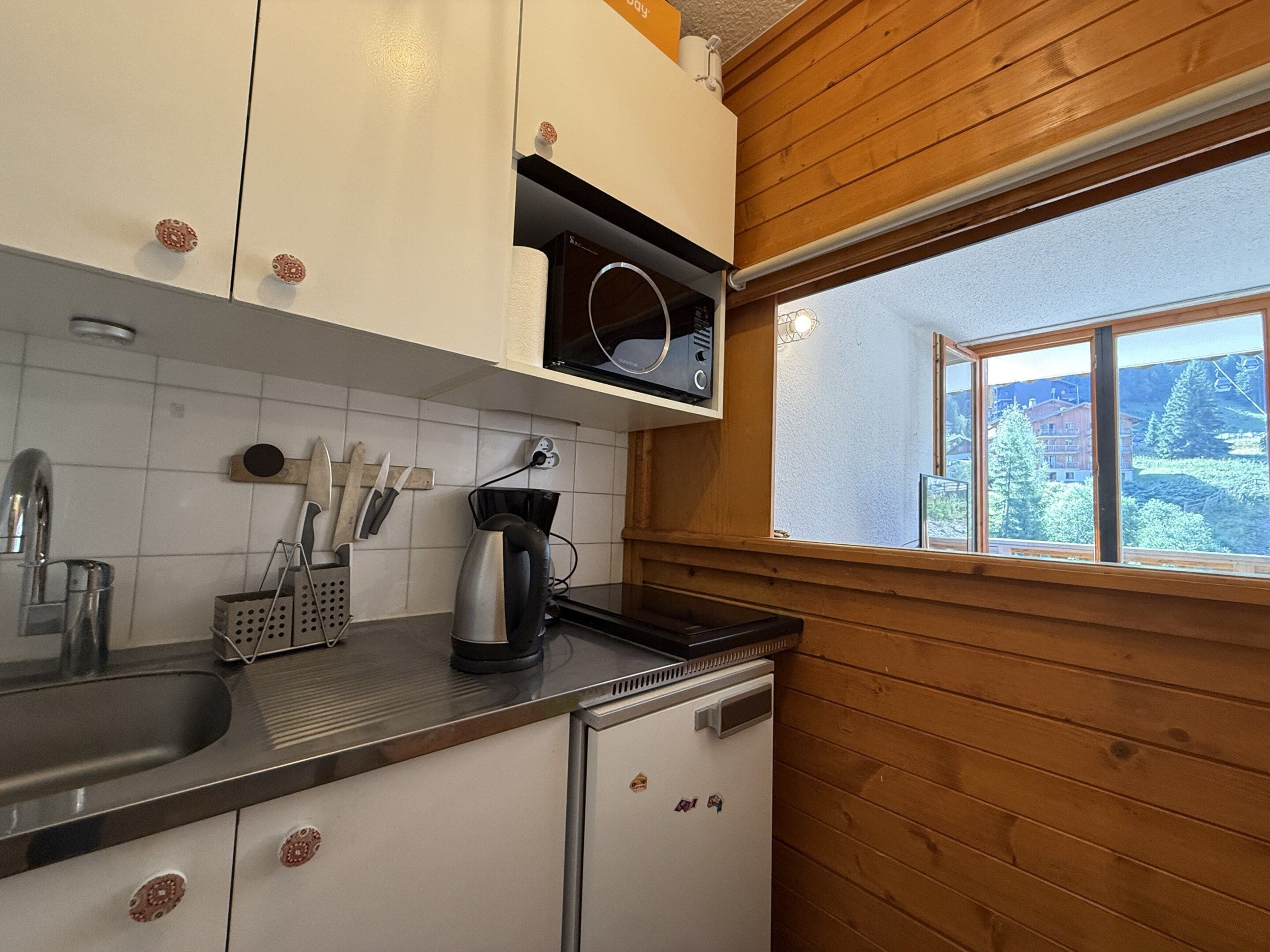 Eine Küche mit weißen Schränken, einer Edelstahl-Spüle und einem Fenster mit Blick auf Bäume und Gebäude.