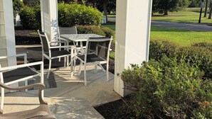 Pátio externo com mesa e cadeiras, cercado por vegetação e céu claro.