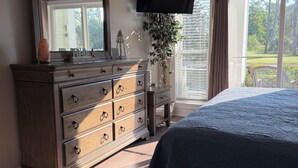 Quarto com cama, cômoda, televisão e janela com vista para área verde.