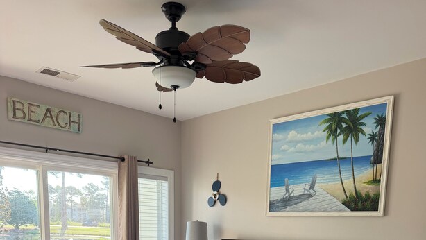 Quarto com cama, ventilador de teto, pintura de uma praia e vista através de uma porta de vidro.