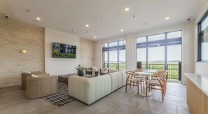 A modern living room with a sectional sofa, a dining area with a round table and chairs, and large windows offering a view of the outdoors. - Modern 2BR near PGA and Universal Studios (Frisco)