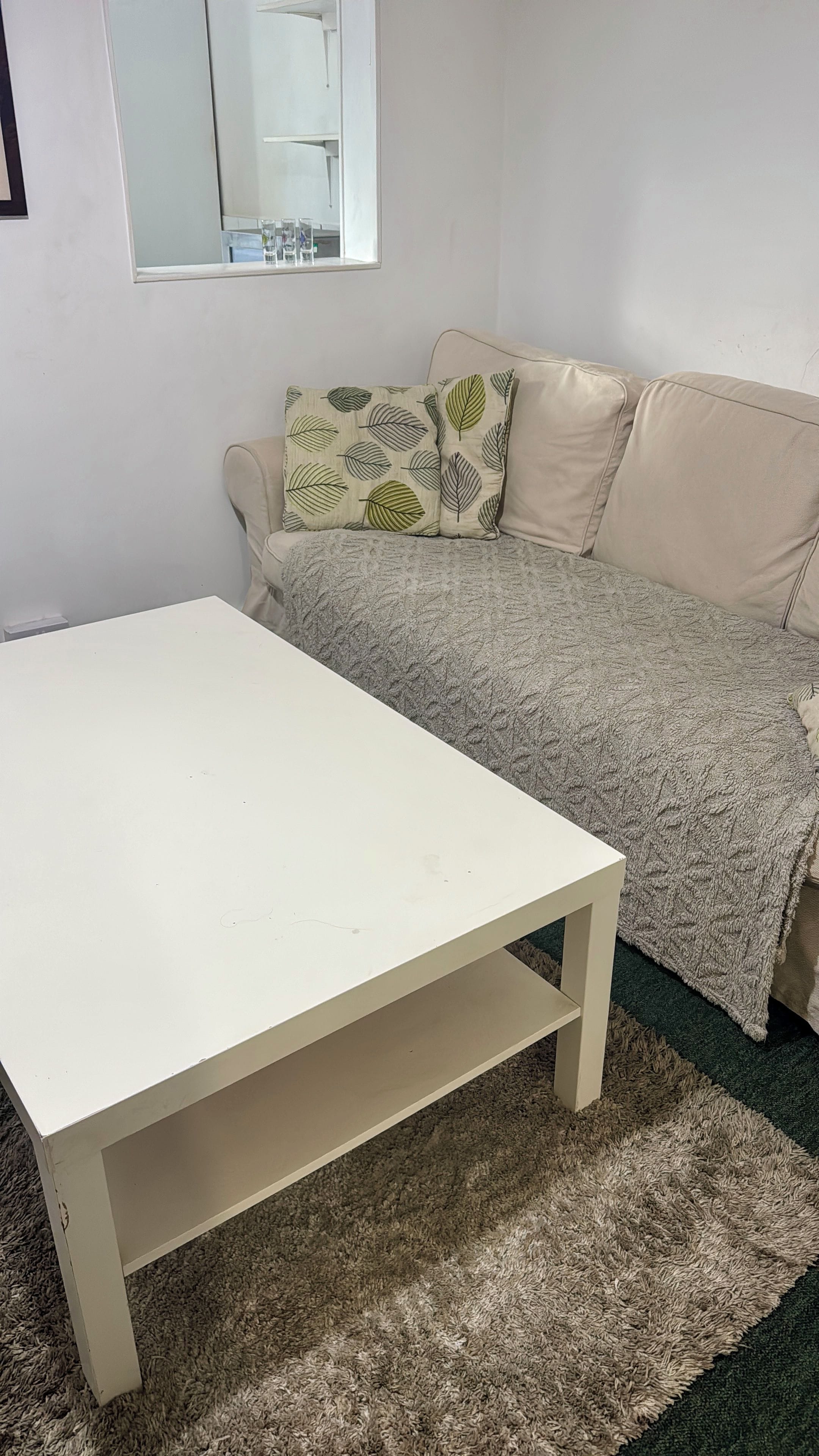 Ein Zimmer mit einem weißen Tisch, einem beigefarbenen Sofa mit gemusterten Kissen und einem Spiegel an der Wand.