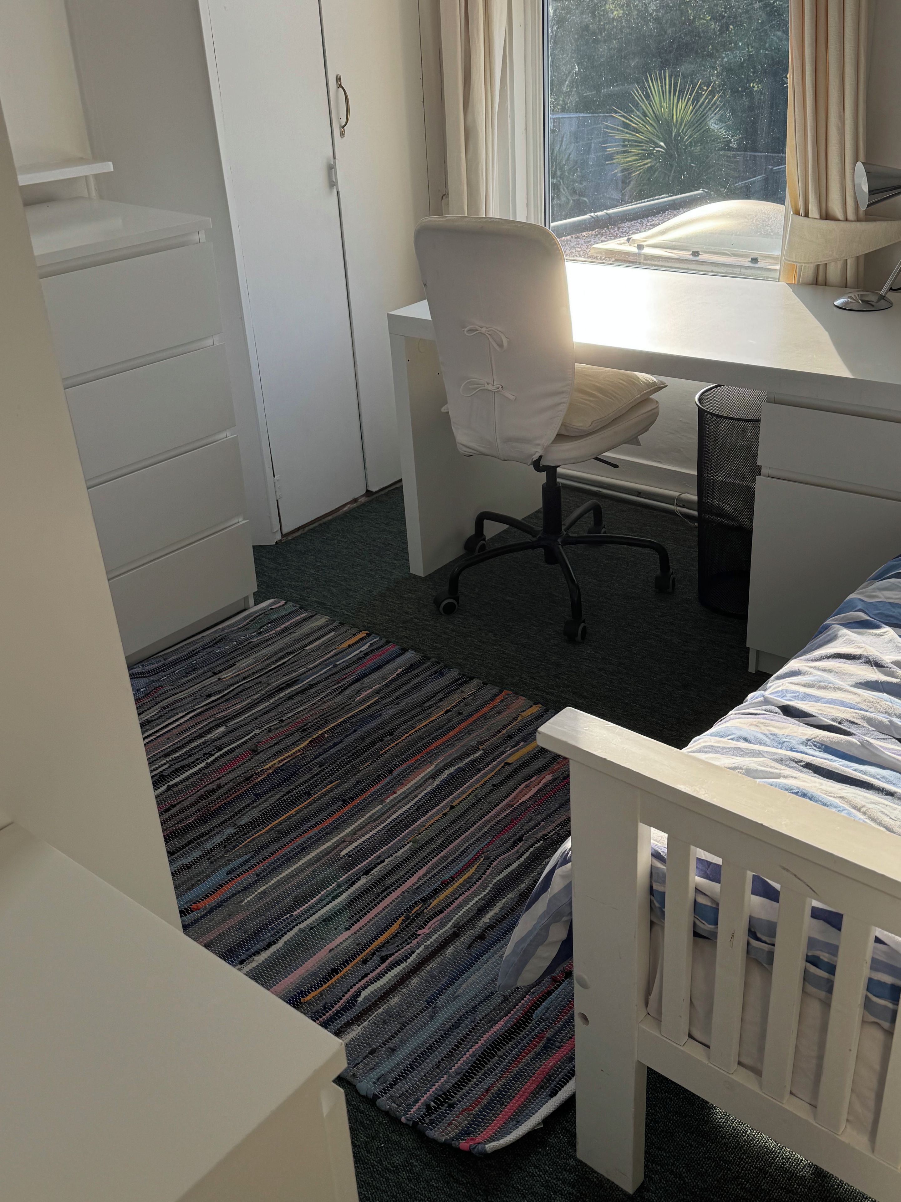 Ein Schlafzimmer mit Bett, Schreibtisch, Stuhl, einem Fenster mit Vorhängen und einem Kleiderschrank.