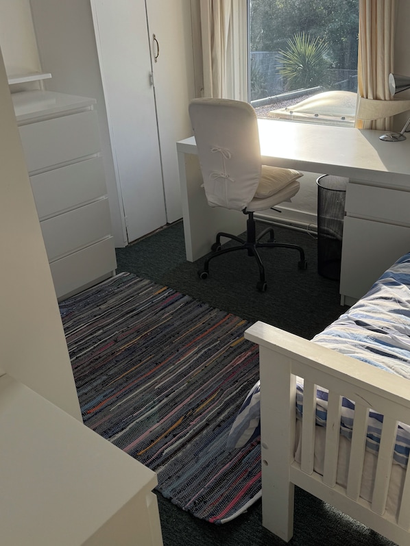 Ein Schlafzimmer mit Bett, Schreibtisch, Stuhl, einem Fenster mit Vorhängen und einem Kleiderschrank.